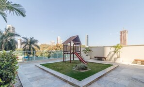A rooftop garden with a children's play structure, including a slide, surrounded by a low stone wall and palm trees.