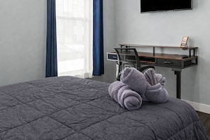 Una cama bien hecha con colcha gris, un escritorio con una silla y un televisor montado en la pared.