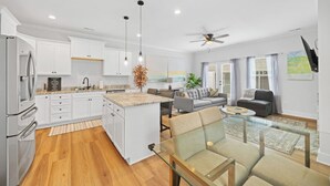 A modern kitchen with white cabinets, a granite countertop, and a central island. There is a dining area with a glass table and chairs, a gray sofa, and a ceiling fan. - New 2024 Townhome | Bright & Modern Stay Close to Downtown Spartanburg (Spartanburg)