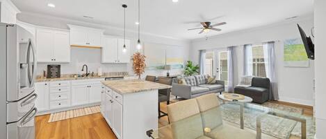 A modern kitchen with white cabinets, a granite countertop, and a central island. There is a dining area with a glass table and chairs, a gray sofa, and a ceiling fan.