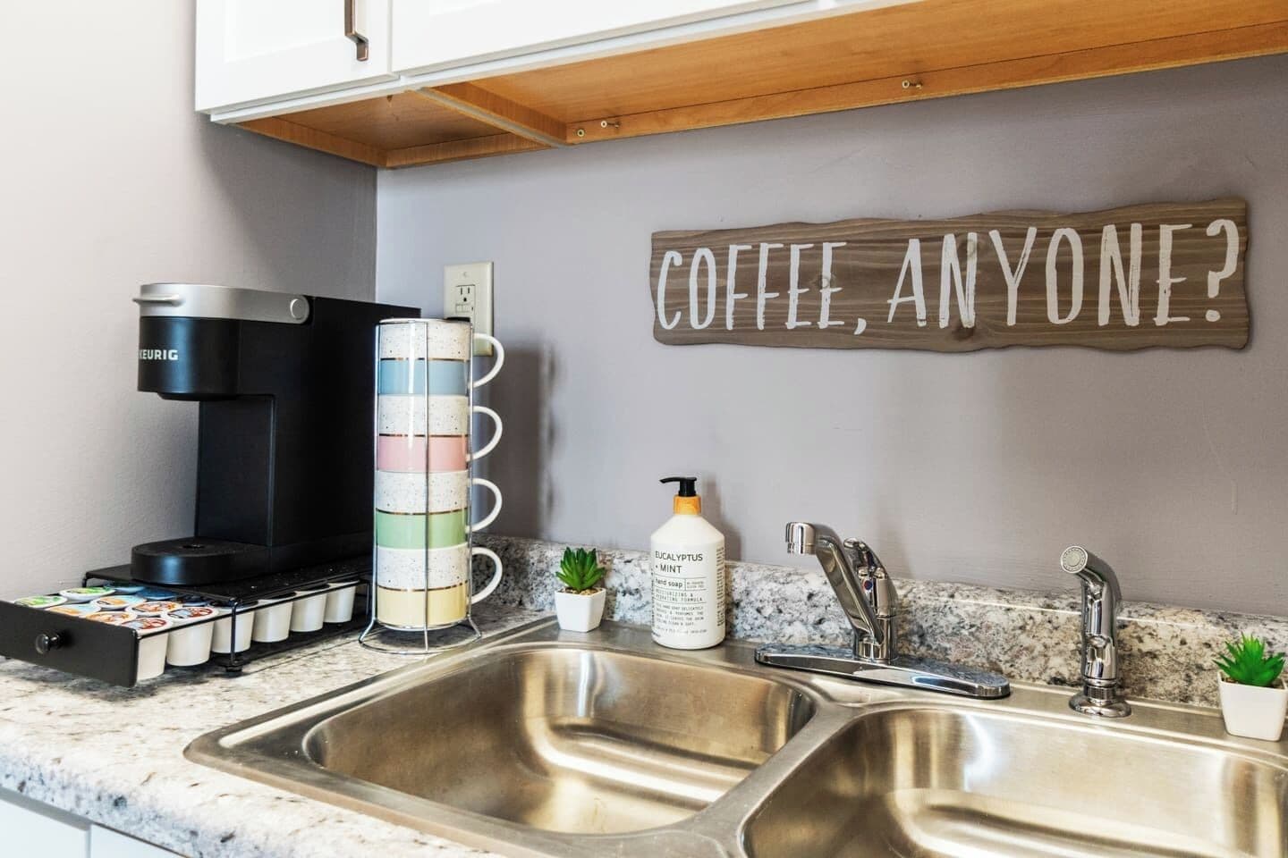 A kitchen with a coffee maker, a mug rack, a bottle of eucalyptus mint soap, and a sign that says "COFFEE, ANYONE?"