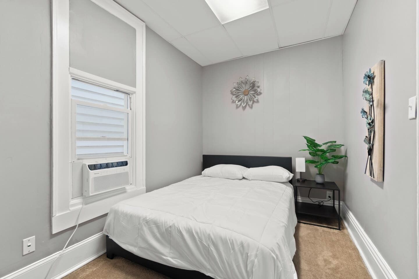 A bedroom with a bed, a nightstand, a wall-mounted flower decoration, and a window with blinds.