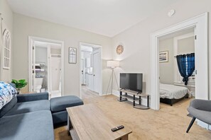 Un salon moderne avec un canapé, une table basse, un téléviseur posé sur un meuble, et une chambre à coucher avec un lit et des rideaux.