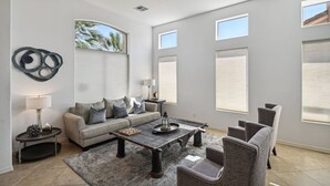 Una sala de estar moderna con un sofá, una mesa de centro y sillas. Hay ventanas amplias y se ve una vista de palmeras.