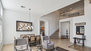 Un salón amplio con una escalera que lleva a un balcón, un comedor con sillas y una mesa, y un letrero que dice 'welcome' en la pared.