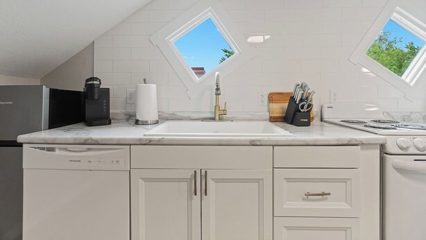 Cocina moderna con armarios blancos, lavavajillas integrado, cafetera, fregadero y estufa. Dos ventanas permiten la entrada de luz natural.