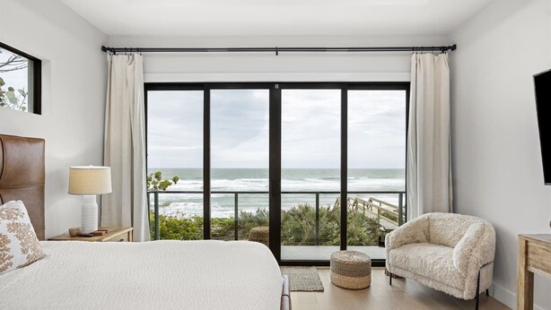 A bedroom with a large window overlooking the ocean, a bed, a chair, and a nightstand with a lamp. - Beachfront Luxe Stay | Pool Access & Rooftop Deck with Stunning Views (Melbourne Beach)