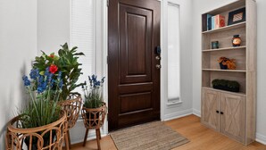 Une porte en bois avec une poignée de porte, un mur blanc, un plancher en bois, une chaise en osier avec une plante, un petit tapis et une bibliothèque en bois contenant des livres et des objets décoratifs.