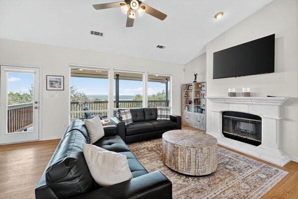 A modern living room with a black leather sectional sofa, a round ottoman, a fireplace, and a large TV mounted on the wall. - Lakeview Luxury with Panoramic views/game room (Lago Vista)