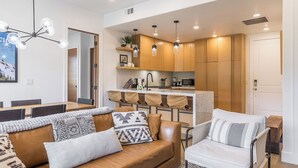 A modern living room with a sofa, a dining table, and a kitchen area with wooden cabinets and pendant lights. - Slope Sight by Avantstay A+ Location w/ Hot Tub (Park City)