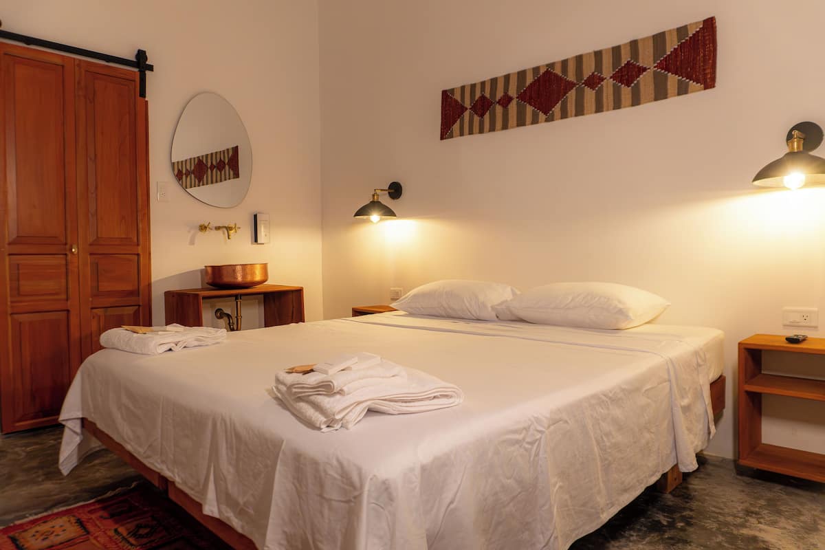 A neatly made bed with white linens, a wooden wardrobe, a mirror, and two wall-mounted lamps.
