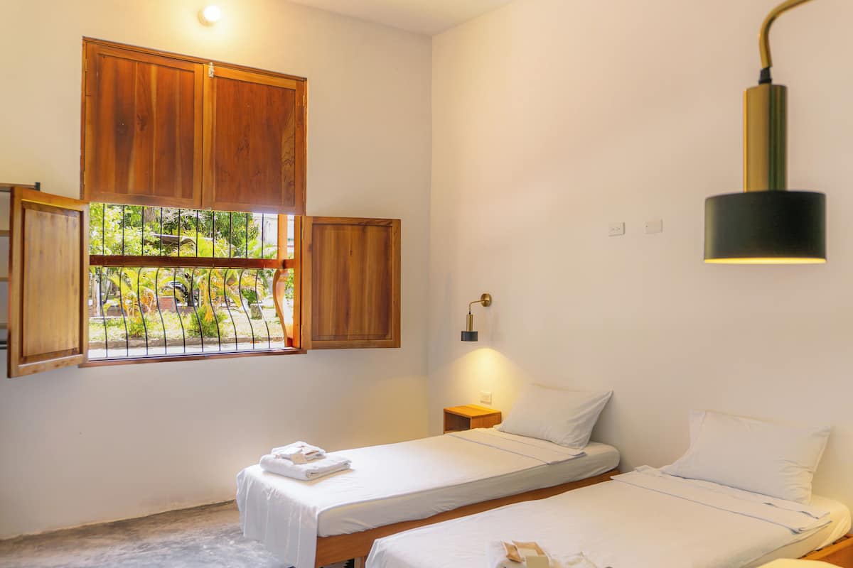 A bedroom with two beds, a window with wooden shutters, a wall-mounted lamp, and a small bedside table.