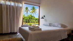 Un dormitorio con cama, ropa de cama blanca, ventana con vista a palmeras, balcón y lámpara fijada a la pared.