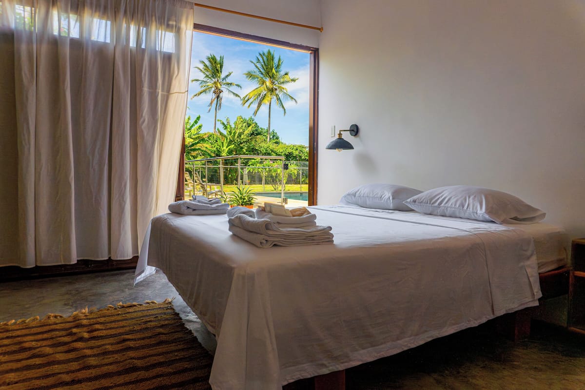 A bedroom with a bed, white linens, a window with a view of palm trees, a balcony, and a wall-mounted lamp.