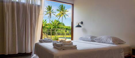 A bedroom with a bed, white linens, a window with a view of palm trees, a balcony, and a wall-mounted lamp.