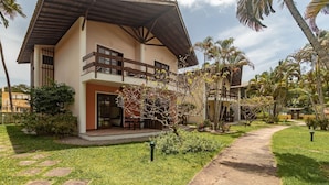 Una casa de dos pisos con balcón, un patio cubierto y un jardín bien cuidado con palmeras y arbustos.