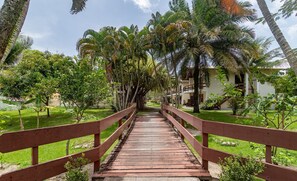 A wooden bridge leading to a building surrounded by lush greenery and palm trees. -  JSO | Conjunto de Apartamentos | Cond. Frente Mar (Porto Seguro)