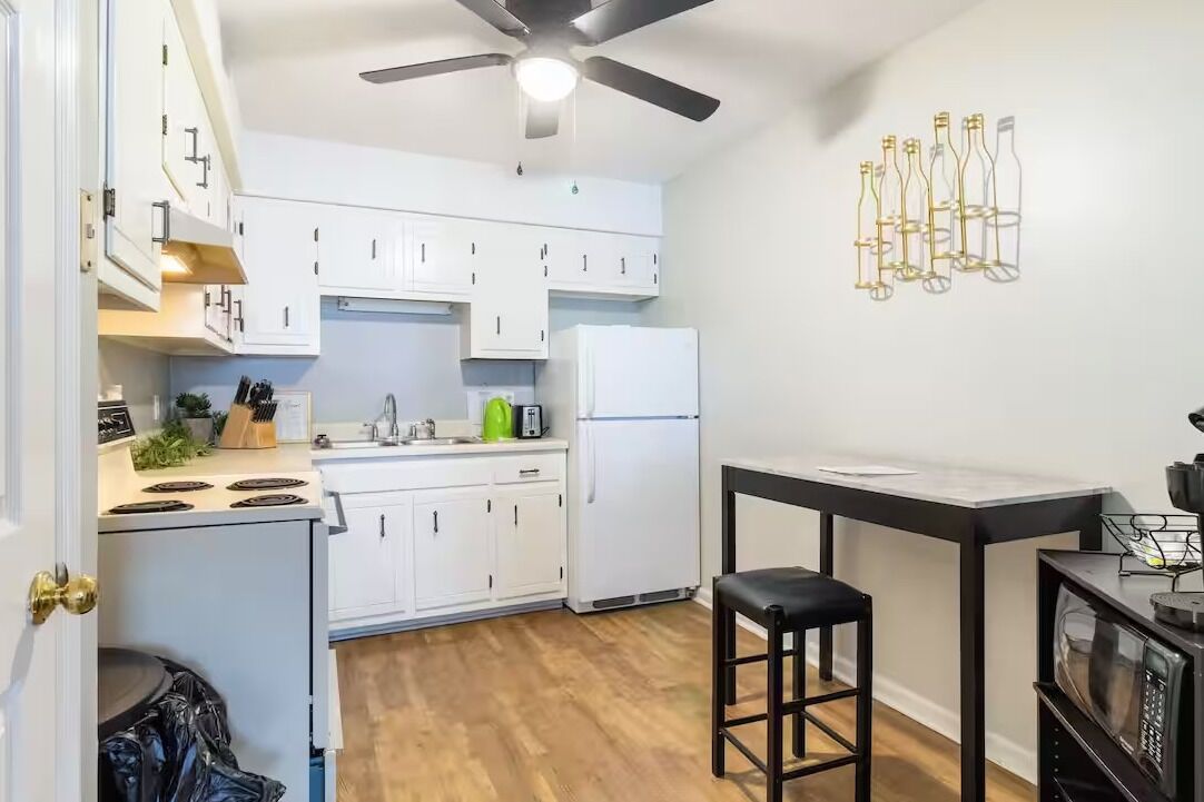 Une cuisine de taille réduite avec des meubles blancs, un ventilateur de plafond noir et un luminaire fixé au mur.
