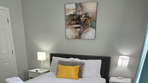 Una cama bien hecha con colcha gris, almohadas blancas y un cojín amarillo decorativo. Hay dos mesitas de noche con lámparas, una alfombra mullida y un cuadro en la pared.