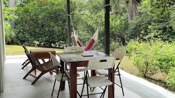 Un espacio de comedor al aire libre techado, con mesa y sillas, rodeado de exuberante vegetación.