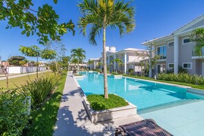 A pool area with a palm tree, a pathway, and a building with multiple windows. - Houses in Condominium Near Pitinga Beach - MGI (Porto Seguro)