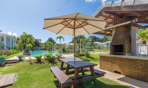 A poolside area with a picnic table and umbrella, surrounded by greenery and a brick barbecue area. - Houses in Condominium Near Pitinga Beach - MGI (Porto Seguro)