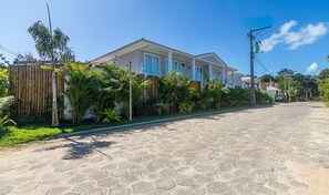 A residential street with a house featuring a tiled roof, a wooden fence, and lush greenery. - Houses in Condominium Near Pitinga Beach - MGI (Porto Seguro)