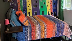 A bedroom with a colorful bedspread, a cityscape mural on the wall, a wooden headboard, a bedside lamp, and a window with curtains. - Miller House Carriage House Studio (mahone bay)