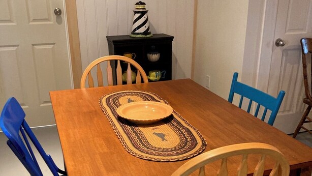 A wooden dining table with a placemat, two chairs, a decorative item on a shelf, and a painting on the wall. - Miller House Carriage House Studio (mahone bay)