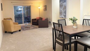 Ein Wohnzimmer mit einer Couch, einem Sessel und einem Esstisch mit Stühlen. Eine Glastür führt auf einen Balkon.