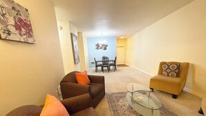 Ein Wohnzimmer mit einer braunen Couch, orangefarbenen Kissen, einem Glastisch und einem gemusterten Teppich. Im Hintergrund befindet sich ein Essbereich mit Tisch und Stühlen.
