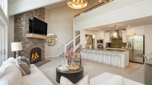 A spacious living room with a stone fireplace, a large TV, and a modern kitchen with stainless steel appliances.
