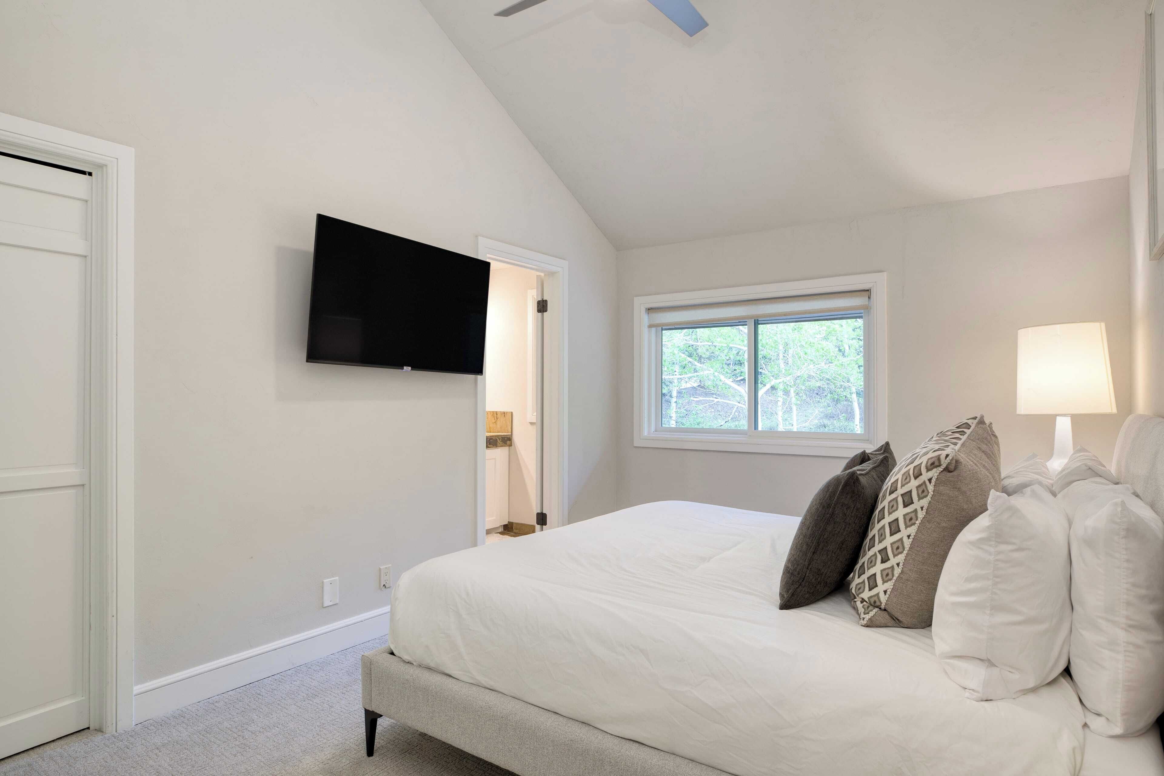 A bedroom with a large bed, a flat-screen TV mounted on the wall, a window with a view of greenery, and a bedside lamp.