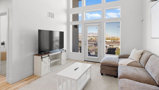 Ein modernes Wohnzimmer mit Flachbildfernseher, weißen Möbeln und großen Fenstern mit Jalousien.