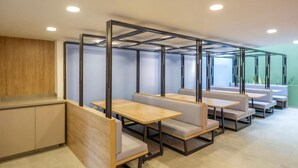 Un área de estar interior moderna y bien iluminada con mesas de madera y bancos acolchados, separada por paneles metálicos.