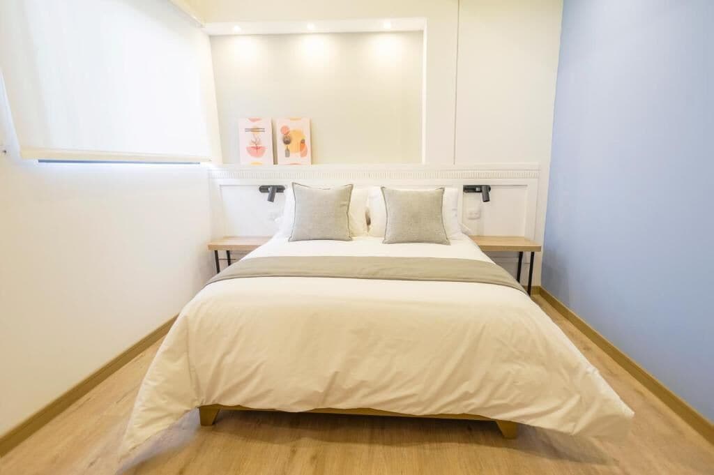 Una cama bien hecha con una colcha blanca y dos almohadas, una mesita de noche con una lámpara, y una estantería de pared con dos cuadros enmarcados.