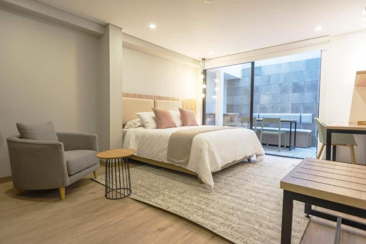 A modern bedroom with a bed, a sofa, a small table, and a balcony with seating.