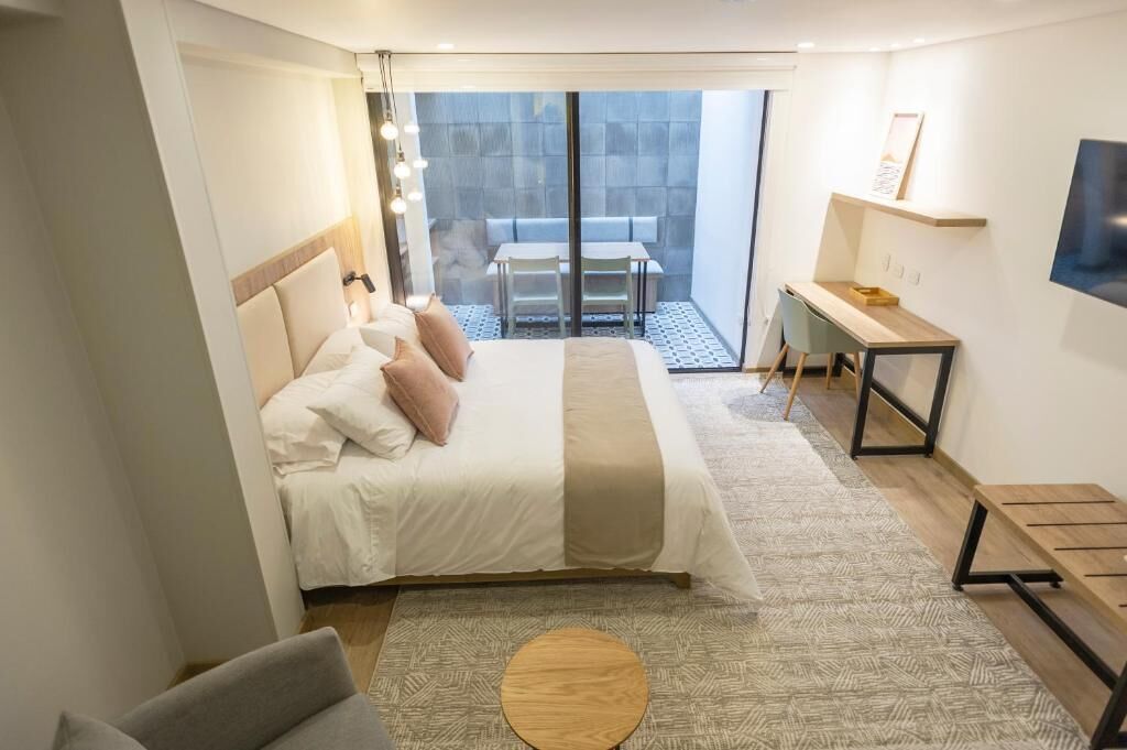 A modern hotel room with a large bed, a desk, a chair, and a balcony with a table and chairs.