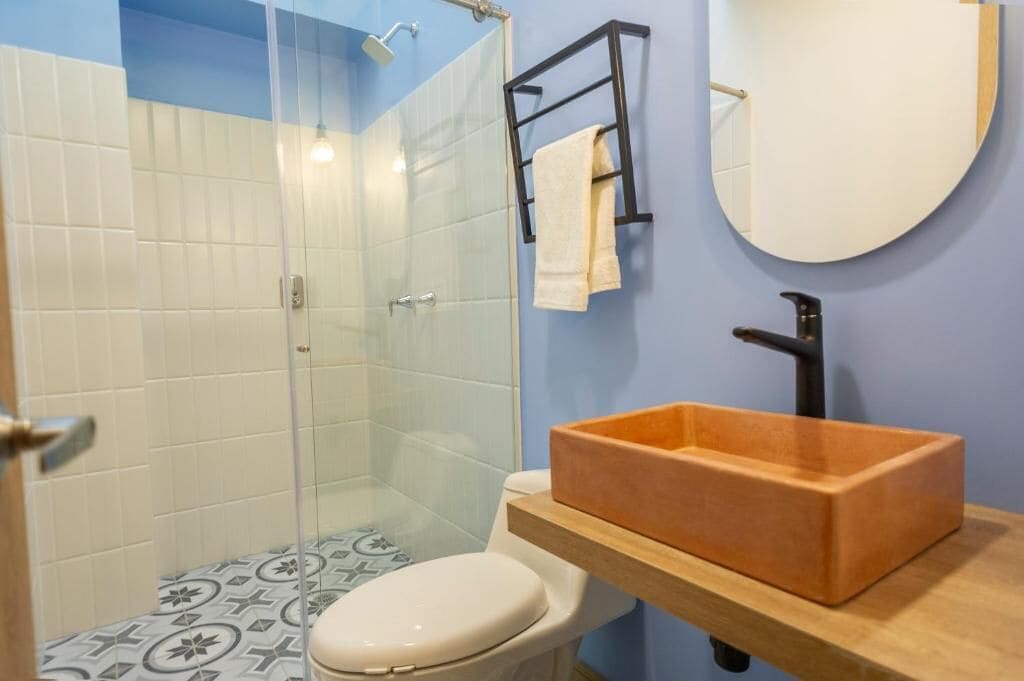 A bathroom with a glass shower enclosure, a white toilet, a wooden vanity with a rectangular sink, and a round mirror.