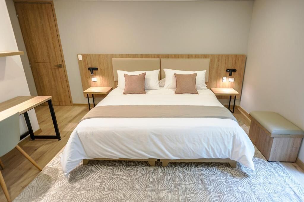 Un dormitorio ordenado con una cama grande, dos mesitas de noche con lámparas, un banco y una mesita auxiliar.
