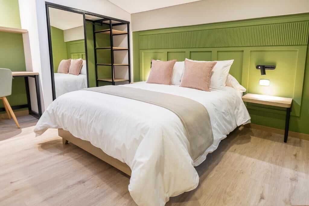 A modern bedroom with a large bed, a green headboard, a bedside table with a lamp, and a wooden floor.