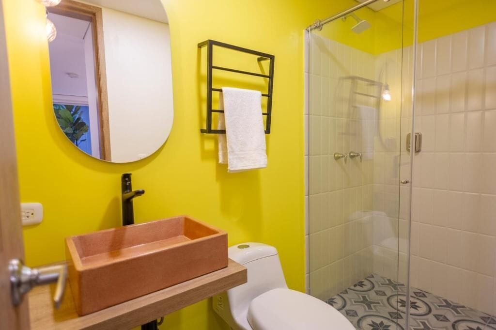 Ein Badezimmer mit gelber Wand, einer weißen Toilette, einem Waschbecken mit Holztablett, einem Spiegel und einer Duschkabine aus Glas.