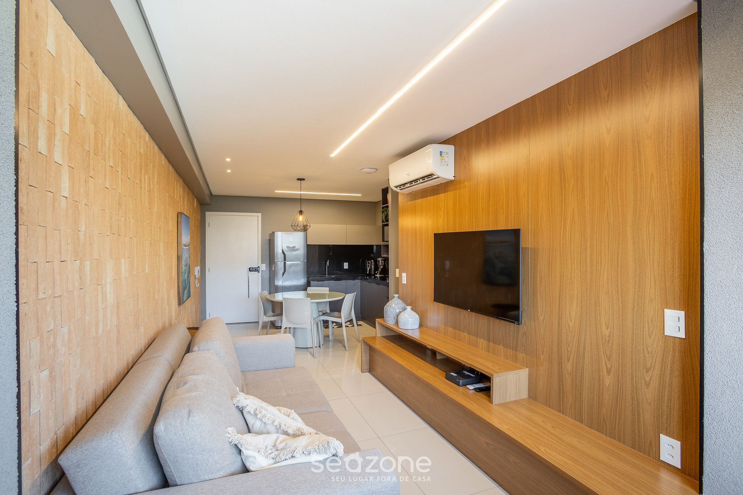 A modern living room with a sofa, a TV mounted on a wooden wall, and a dining area in the background.