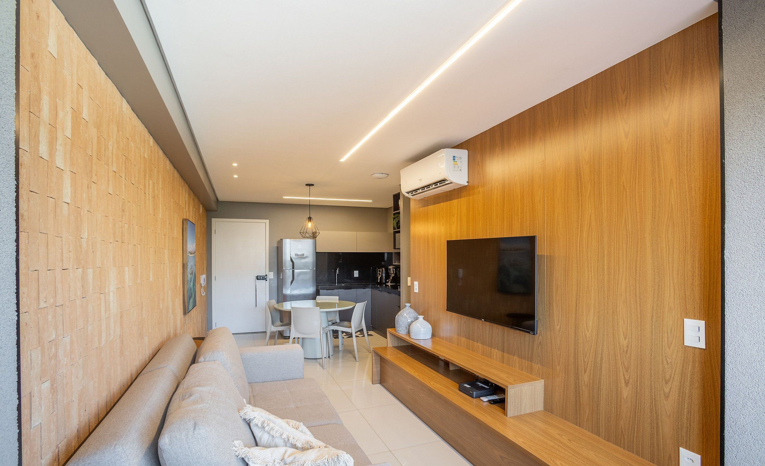 Uma sala de estar moderna com um sofá, uma TV fixada em uma parede de madeira, e uma área de jantar ao fundo.