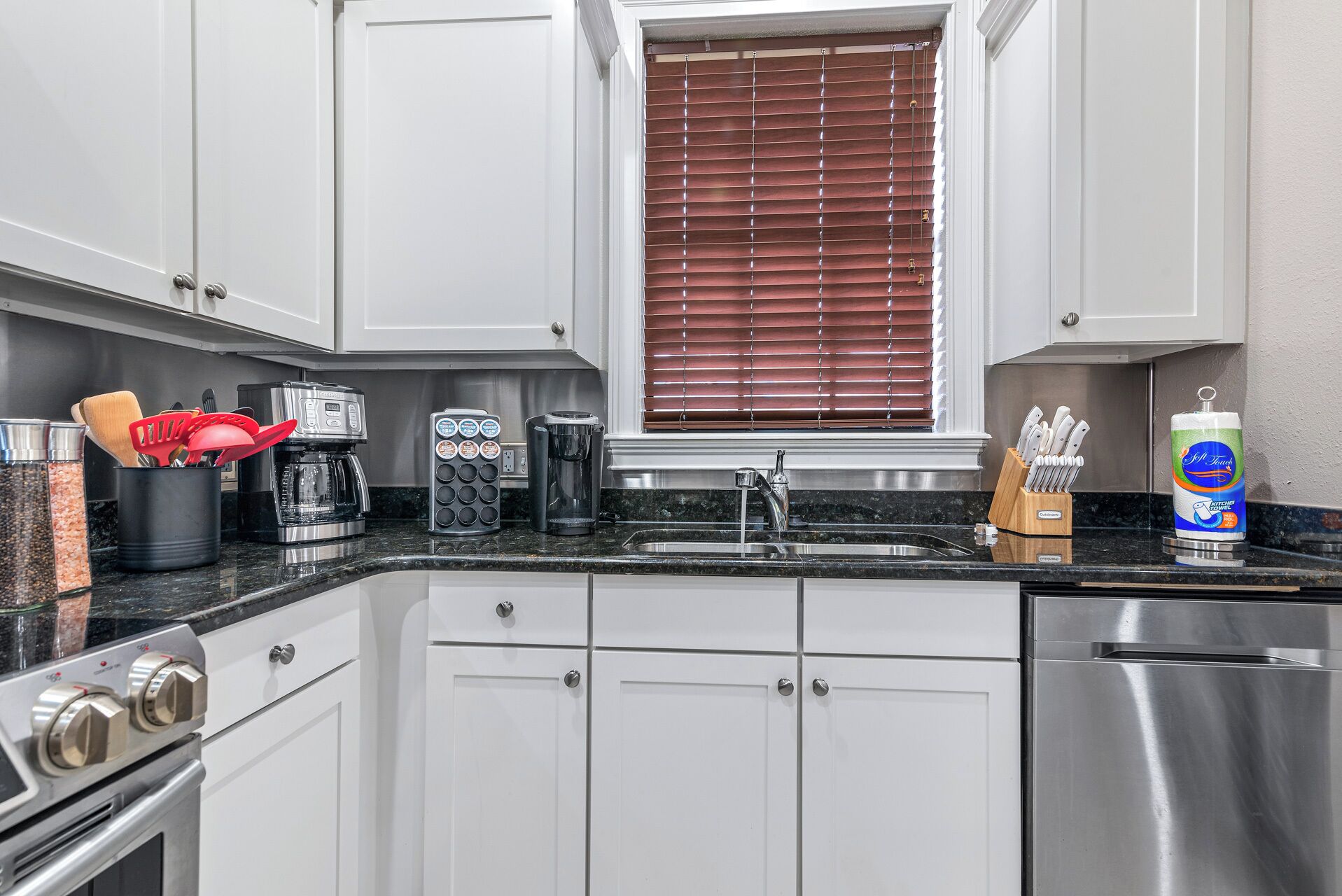 A modern kitchen with white cabinets, stainless steel appliances, and a window with brown blinds.