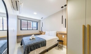 Ein modernes Hotelzimmer mit einem großen Bett, einem kleinen Tisch, einem Fenster mit Blick auf Gebäude und einer Decken-Klimaanlage.