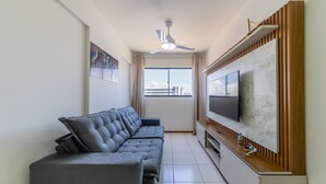 Een moderne woonkamer met een grijze bank, een plafondventilator, een aan een houten paneel bevestigde televisie en een raam met uitzicht op gebouwen.