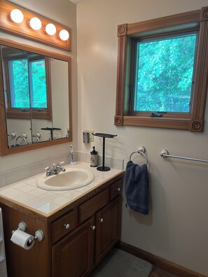 A bathroom with a wooden vanity, a sink, a mirror with lights, a window with a view of greenery, and a towel rack. - Chippewa Bay Retreat (Hammond)