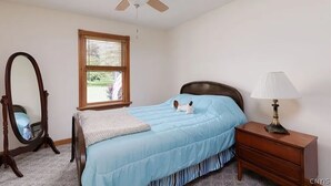 A bedroom with a bed, a nightstand, a lamp, a ceiling fan, a window, and a mirror. - Chippewa Bay Retreat (Hammond)