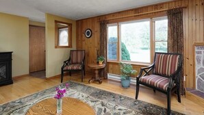 A spacious living room with wooden walls, a ceiling fan, two armchairs, a coffee table, and a large window with curtains. - Chippewa Bay Retreat (Hammond)